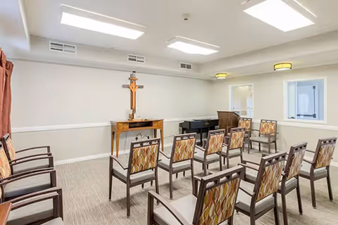 Small chapel-style meeting room with rows of patterned chairs facing a wooden cross on a table and a piano.
