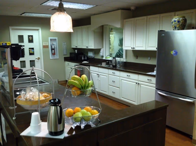 Interior view of a kitchen area with white cabinets, a stainless steel refrigerator, a countertop with a fruit basket containing bananas, oranges, and apples, a coffee pot with disposable cups, and a hanging light fixture above the counter.