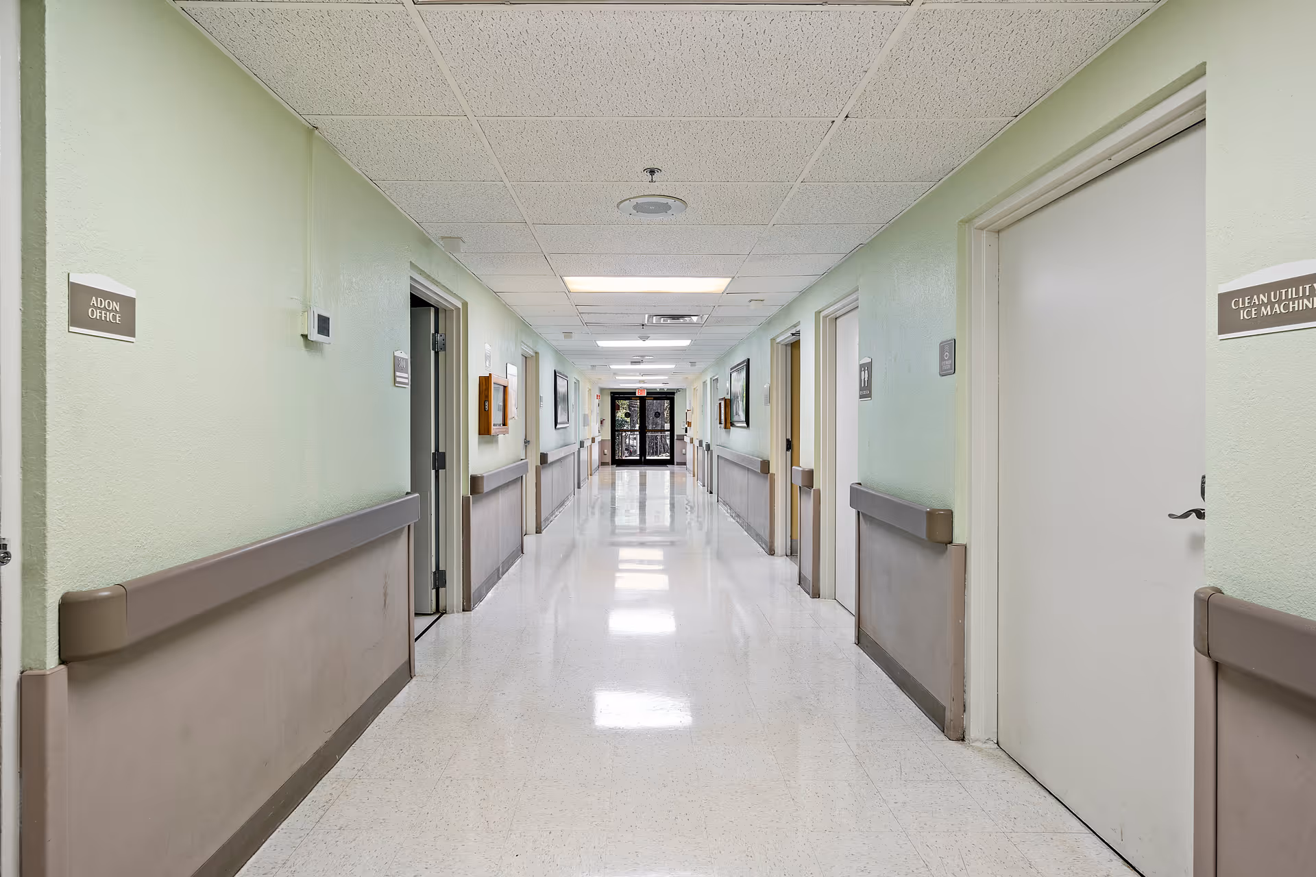A clean, well-lit hallway in a senior living facility with light green walls, beige handrails, and white tiled floor. Several closed doors line both sides of the corridor, with signs indicating rooms such as 'ADON OFFICE' and 'CLEAN UTILITY ICE MACHINE'. The hallway extends to a glass exit door at the far end.