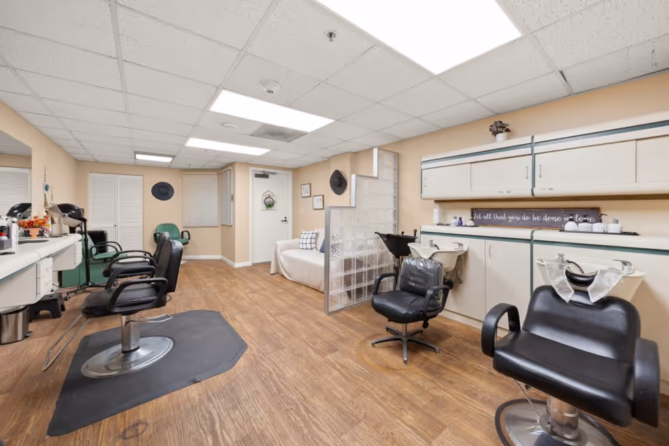 Interior of a hair salon area in a senior living facility with multiple black salon chairs, hair washing stations, a white couch, and beige walls with decorative items including hats and framed pictures.