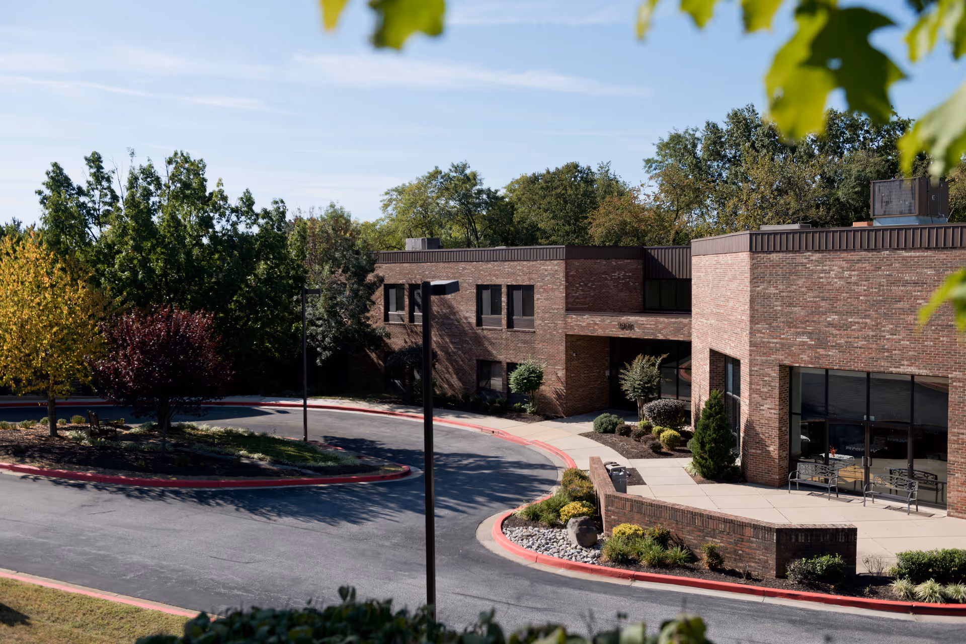Brick senior living facility entrance with a curved driveway, landscaped islands, and surrounding trees.