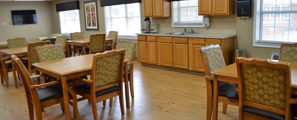 A dining room with multiple wooden tables and chairs with green patterned upholstery. The room has large windows with blinds, a wall-mounted television, and a kitchenette area with wooden cabinets, a sink, and a paper towel dispenser. The floor is light wood.
