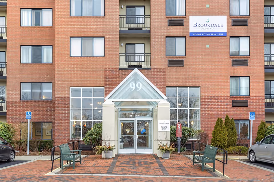 Front entrance of a brick Brookdale Quincy Bay senior living building with a glass canopy marked 99, benches, and signage.