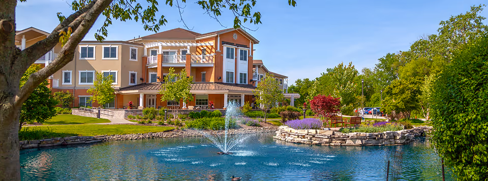 A large assisted living facility building with multiple stories, surrounded by well-maintained landscaping including trees, shrubs, and colorful flowers. In the foreground, there is a pond with a water fountain spraying water upwards, and a stone-bordered garden area with benches. The sky is clear and blue.