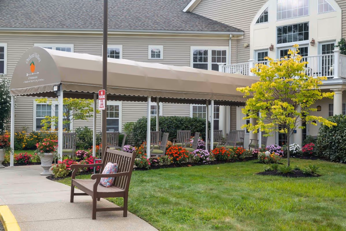 Outdoor seating area at Benchmark at Orchard Valley with a covered walkway, wooden benches with cushions, colorful flower beds, a small tree with yellow leaves, and a beige building with multiple windows in the background.