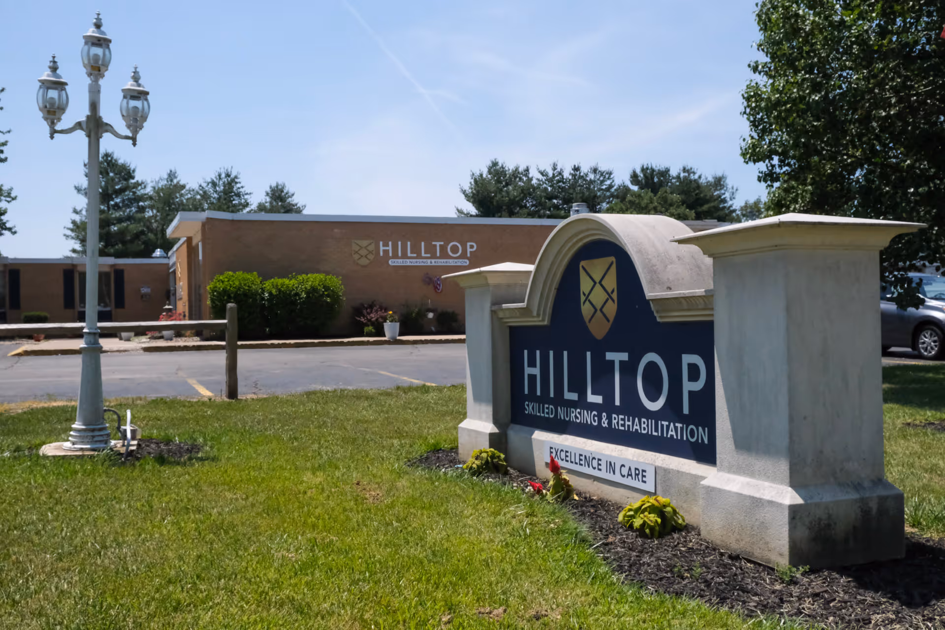 A stone entrance sign reading 'Hilltop Skilled Nursing & Rehabilitation' on a grassy lawn with the facility building, a lamppost, and parked car in the background.