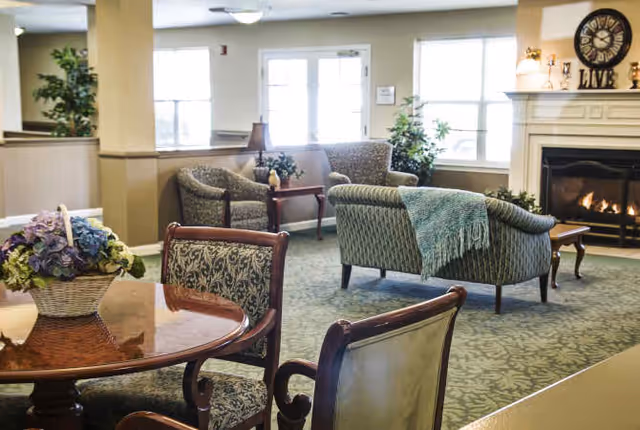 A cozy senior living facility common area with patterned armchairs and a loveseat arranged around a fireplace. A wooden table with floral centerpiece is in the foreground, and large windows allow natural light to fill the room.