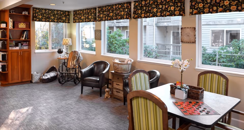 A cozy common area in a senior living facility with large windows letting in natural light. The room features a table with four chairs, a checkers game set up on the table, two leather armchairs, a small wooden cabinet with baskets, a rocking chair with a throw blanket, a floor lamp, and a wooden bookshelf filled with books and games. The windows have floral valances, and outside greenery and neighboring buildings are visible.