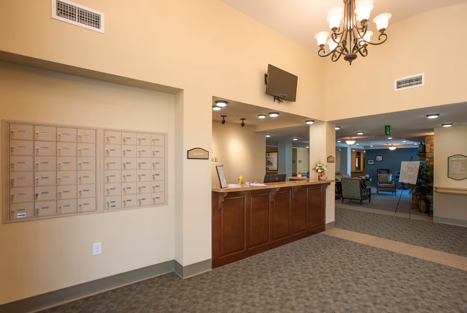 Interior view of a senior living facility reception area with a wooden front desk, a wall-mounted TV above the desk, and a set of mailboxes on the left wall. The area is well-lit with ceiling lights and a chandelier, and there is a seating area visible in the background.