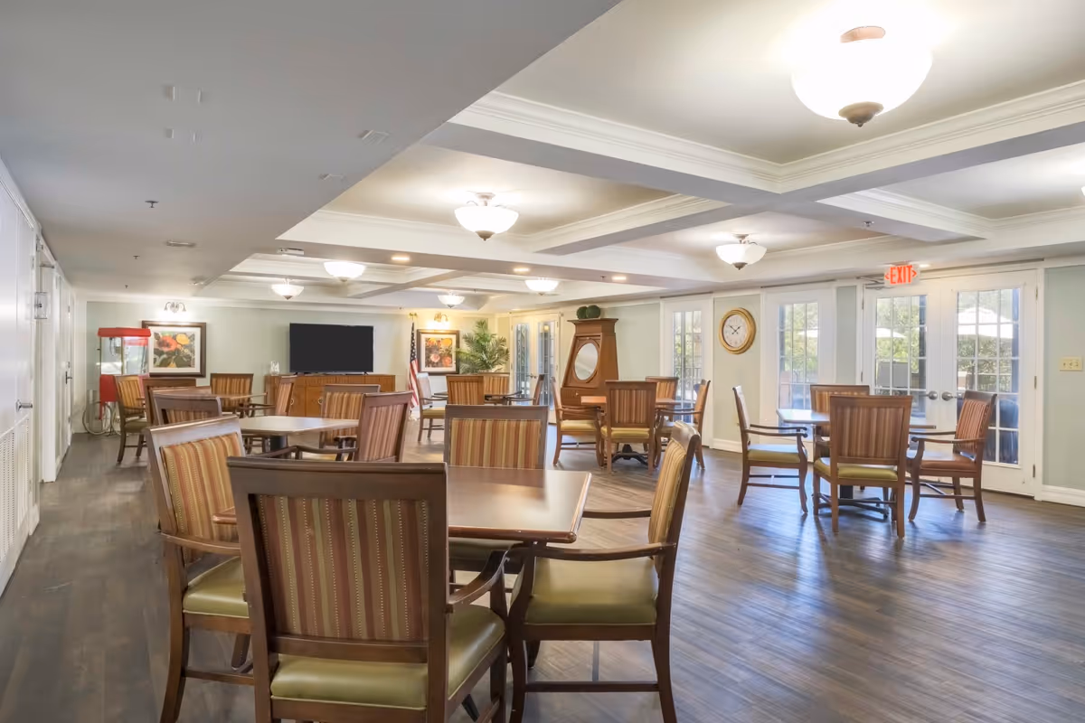 Bright common dining room with several wooden tables and upholstered chairs, a TV, clock, and glass doors to the exterior.