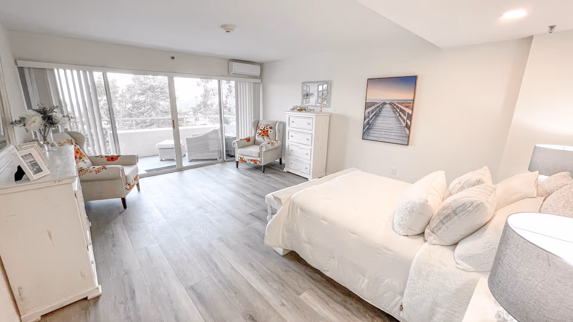 A bright and spacious bedroom with a large bed covered in white bedding and multiple pillows. The room features two floral upholstered armchairs near a sliding glass door that leads to a balcony with outdoor seating. There is a white dresser with a mirror and a framed picture of a boardwalk on the wall. The floor is light-colored wood, and the walls are painted white.