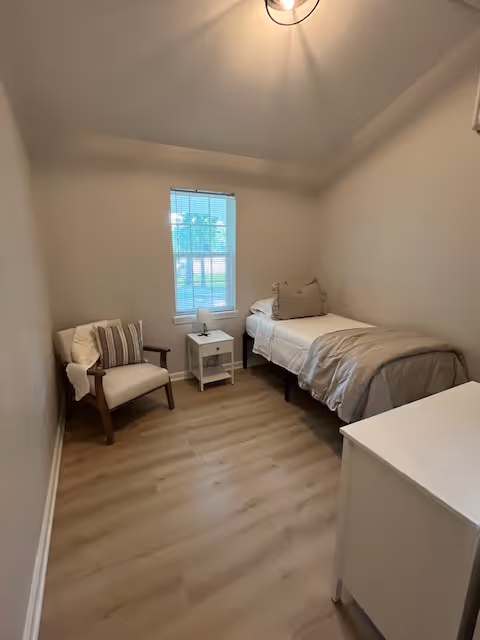 A small, simple bedroom with a single bed against the right wall, a white nightstand with a lamp next to the bed, a cushioned armchair with a striped pillow on the left side, a window with blinds letting in natural light, and light wood flooring.