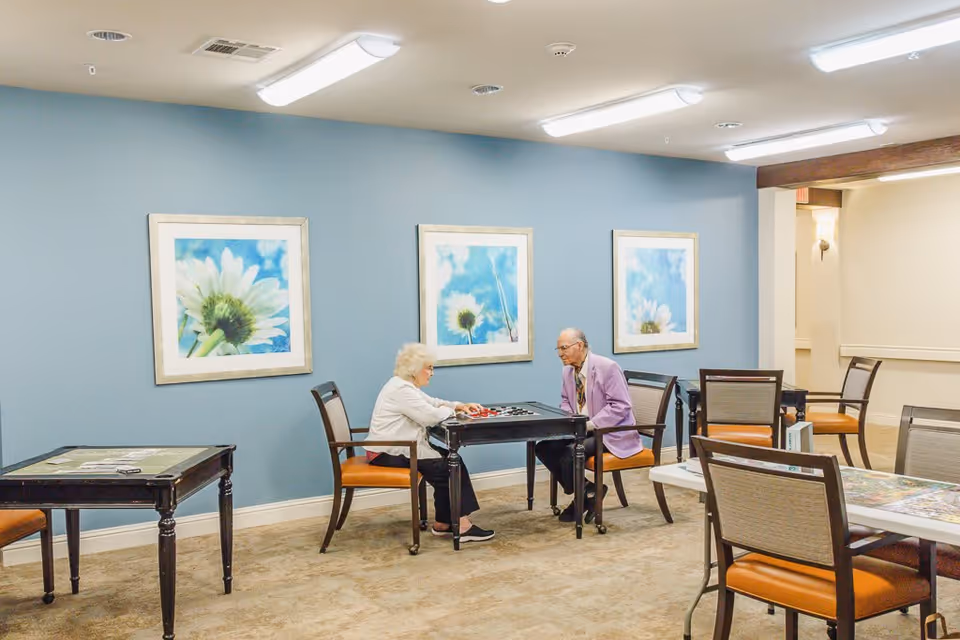 Two elderly individuals sitting across from each other at a small table playing a board game in a well-lit room with blue walls adorned with three framed pictures of white flowers against a blue sky. The room has several other tables and chairs arranged around it.