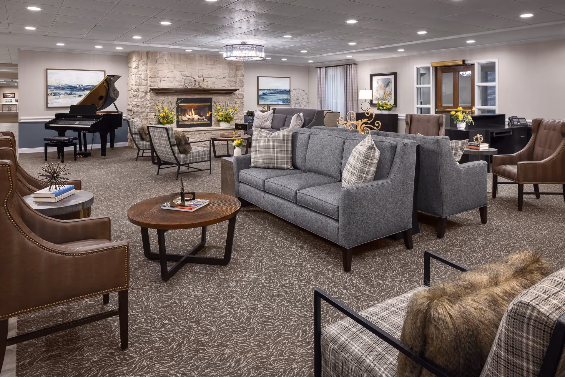 A spacious and elegantly furnished senior living common area featuring gray sofas with plaid pillows, brown leather armchairs, a round wooden coffee table, and a stone fireplace with a fire burning. A grand piano is positioned in the corner, and the room is decorated with framed artwork, flowers, and soft lighting from ceiling fixtures and lamps.