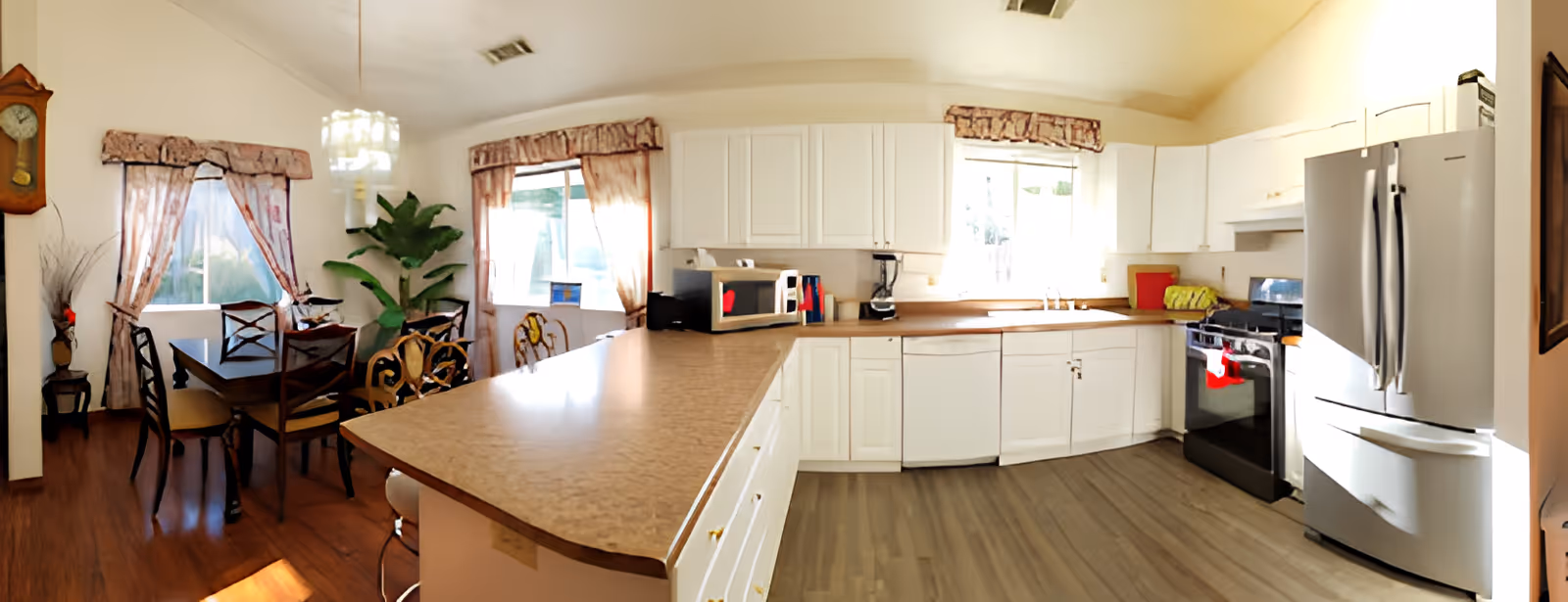 Bright open kitchen with white cabinets, a long countertop island, stainless refrigerator and stove, and a dining table with chairs by curtained windows.