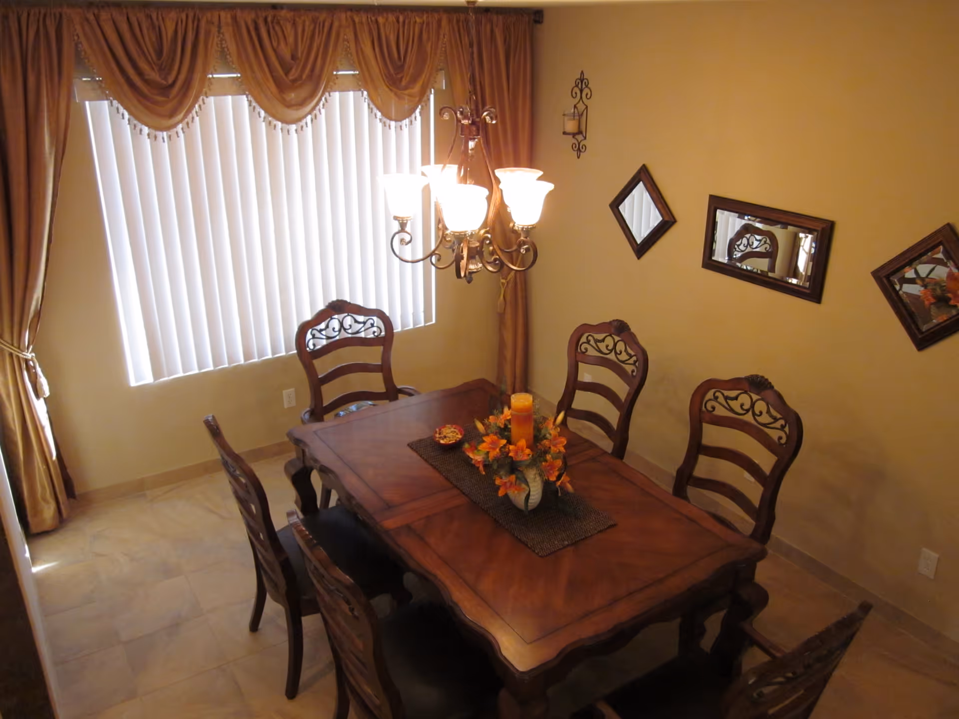 A dining room with a wooden dining table surrounded by six matching chairs. The table has a centerpiece with orange flowers and a candle. There is a chandelier with five lights hanging above the table. The room has beige walls with three decorative mirrors and a wall-mounted candle holder. A large window with vertical blinds and brown curtains is on one side of the room.
