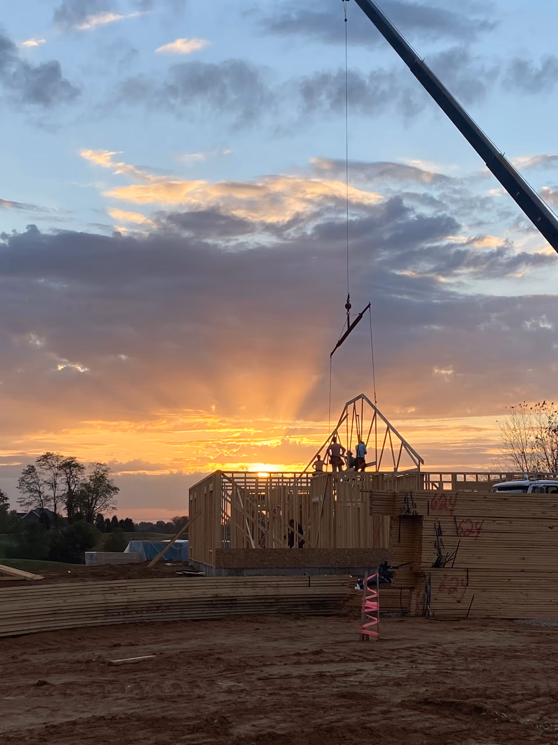 Sunset over a construction site with workers and a crane lifting a roof truss onto a wooden-framed building.