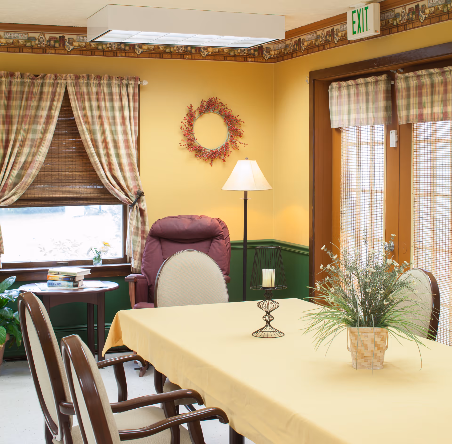 A cozy dining/activity room with a yellow tablecloth-covered table and chairs, a potted plant centerpiece, plaid curtains, a reclining chair and floor lamp.
