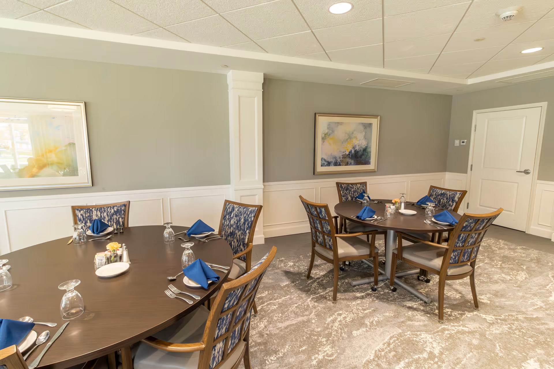 A dining room with two round tables set for meals, each with blue napkins, plates, silverware, and glasses. The room has light gray walls with white wainscoting, two framed abstract paintings, a patterned carpet, and a white door.
