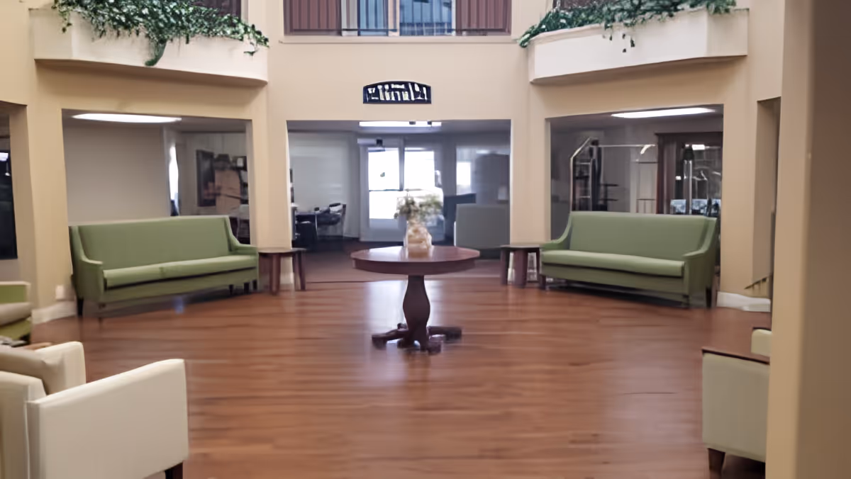 A spacious interior common area with wooden flooring, featuring two green sofas against the walls and two white armchairs in the foreground. A round wooden table with a flower vase is centered in the room. There are large windows and doorways in the background, and some greenery decorates the upper walls.