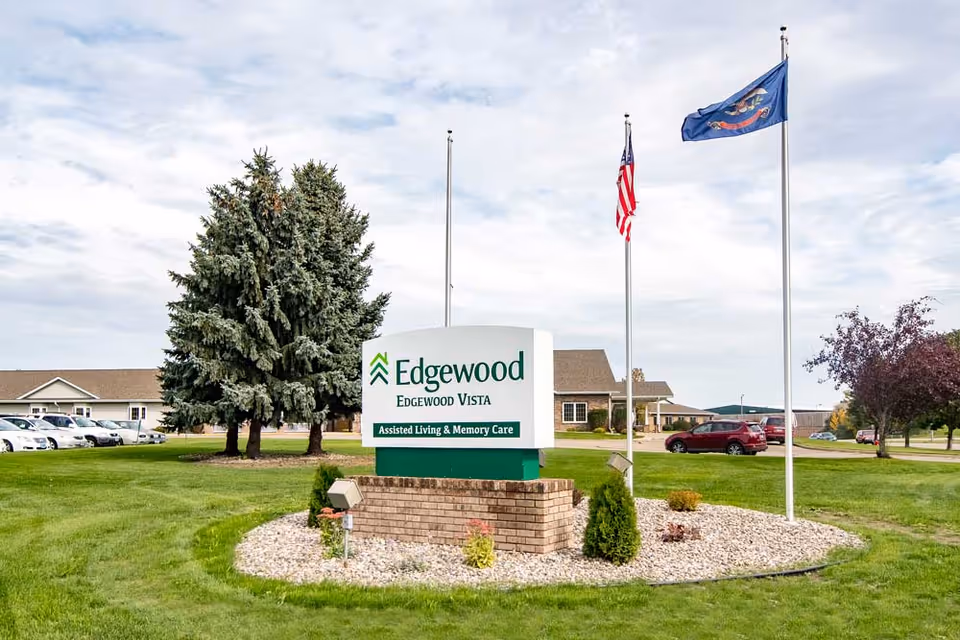 Outdoor view of the Edgewood Vista assisted living and memory care facility sign on a grassy area with trees, parked cars, and two flagpoles displaying the American flag and another flag.