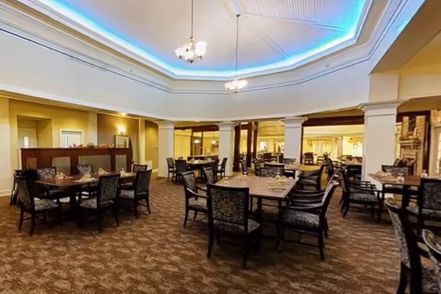 A spacious dining room with multiple wooden tables and cushioned chairs arranged neatly. The ceiling features a recessed design with blue accent lighting and two chandeliers. The room has beige walls and carpeted flooring with a patterned design.