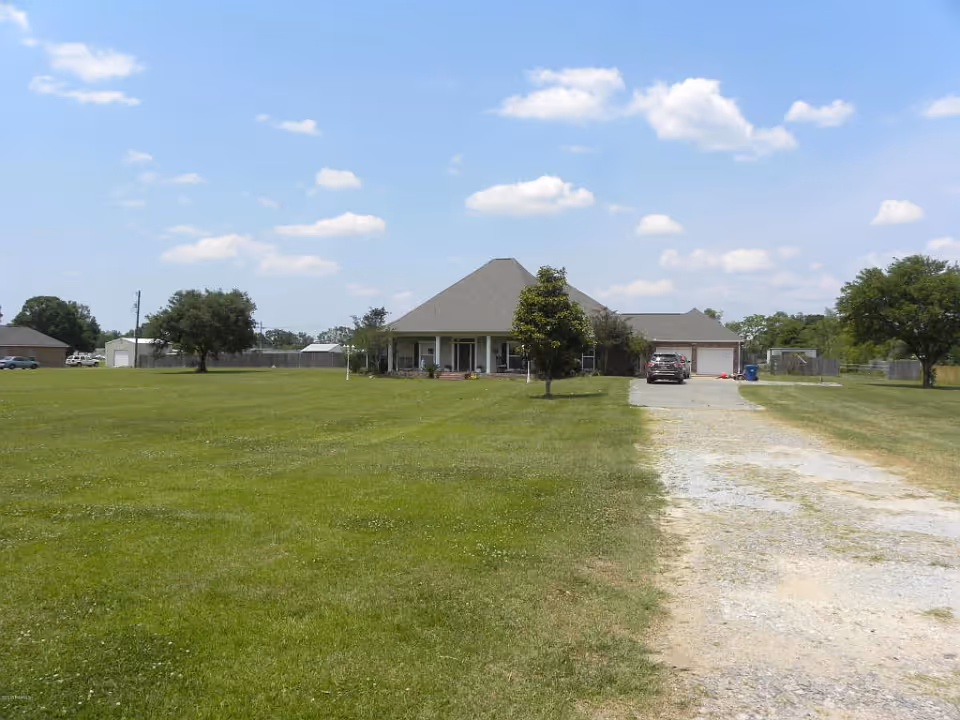 Single-story house with a large grassy front lawn and a gravel driveway leading to a car and garage under a partly cloudy sky.