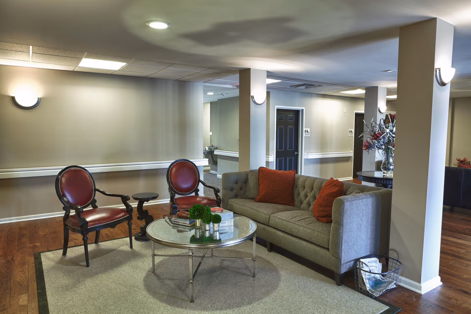 A cozy seating area in a senior living facility with a gray tufted sofa adorned with red cushions, two red leather armchairs, and a round glass coffee table with decorative items. The room has wooden flooring, beige walls, and soft lighting from wall sconces. In the background, there are doors and a hallway leading to other parts of the facility.