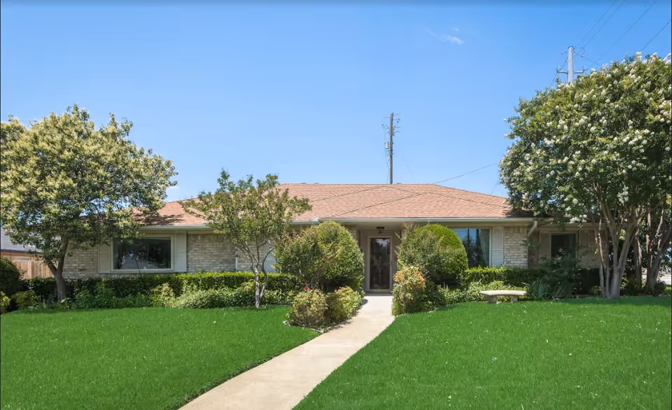 Single-story brick building with a brown shingled roof, surrounded by green grass, bushes, and several trees under a clear blue sky. A concrete walkway leads to the front door.