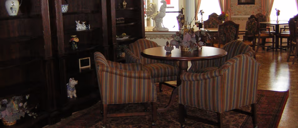 A cozy interior room with a round wooden table surrounded by four striped upholstered chairs. Behind the table is a dark wooden bookshelf filled with decorative items and figurines. The room has a warm ambiance with patterned rugs and additional seating visible in the background near windows with curtains.