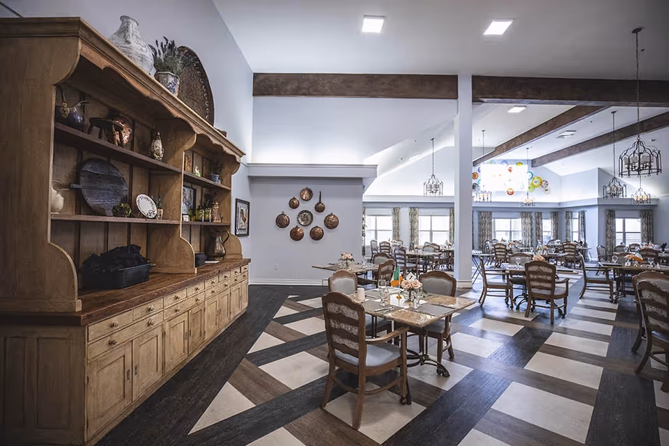 A spacious dining room with multiple tables and chairs arranged neatly. The room features high ceilings with exposed wooden beams and decorative hanging light fixtures. On the left side, there is a large wooden cabinet with shelves displaying various decorative items. The floor has a patterned design with dark and light wood tones. The walls are light-colored with windows allowing natural light to enter, and decorative plates are hung on one wall.