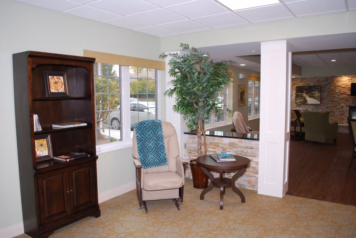 A cozy seating area with a recliner, round side table, bookshelf, and large potted plant beside windows.