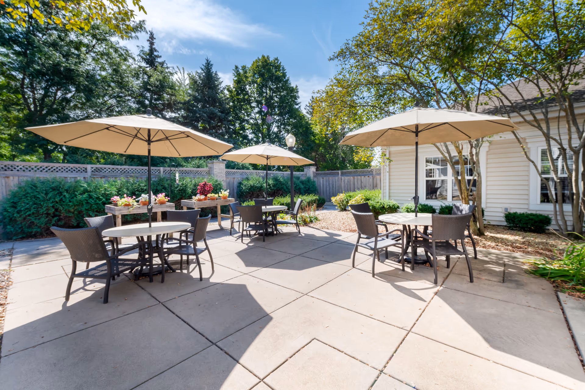 Outdoor patio area with round tables and chairs under large beige umbrellas, surrounded by greenery and a wooden fence, with a building visible on the right side.