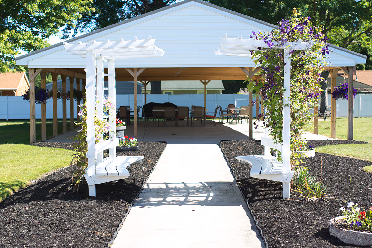 Outdoor garden area with a concrete pathway leading to a covered pavilion. Two white wooden trellises with climbing purple flowers and attached benches flank the pathway. The pavilion has several tables and chairs underneath, surrounded by green grass and trees.