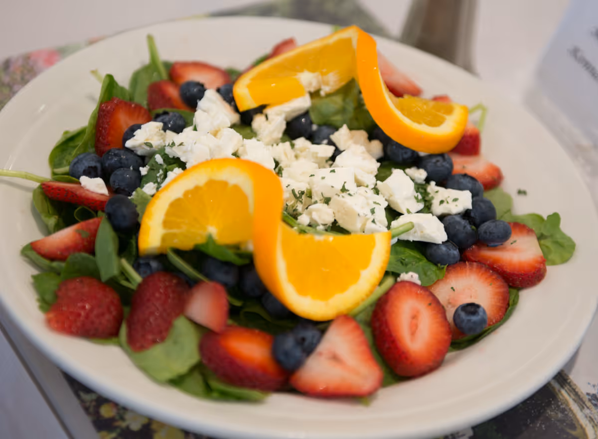 A fresh salad on a white plate featuring spinach leaves, blueberries, sliced strawberries, crumbled white cheese, and two orange slices on top.