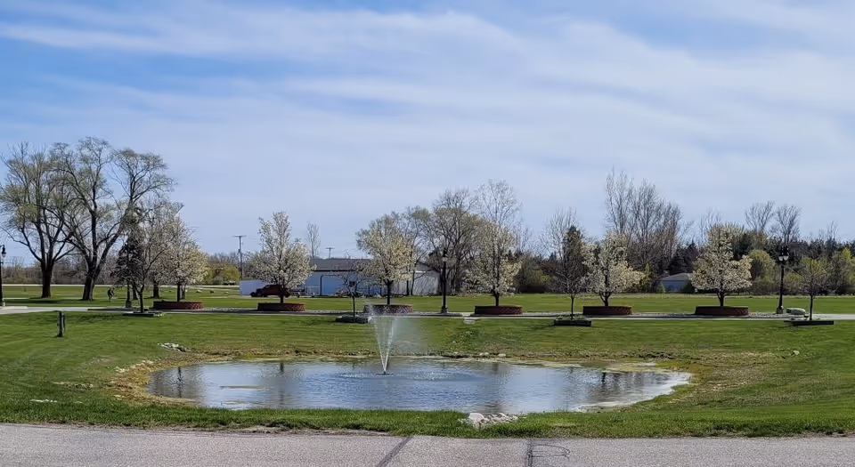 A small pond with a water fountain in the center, surrounded by green grass and a row of trees with light-colored blossoms. There is a paved road in the foreground and a clear blue sky with some clouds above.