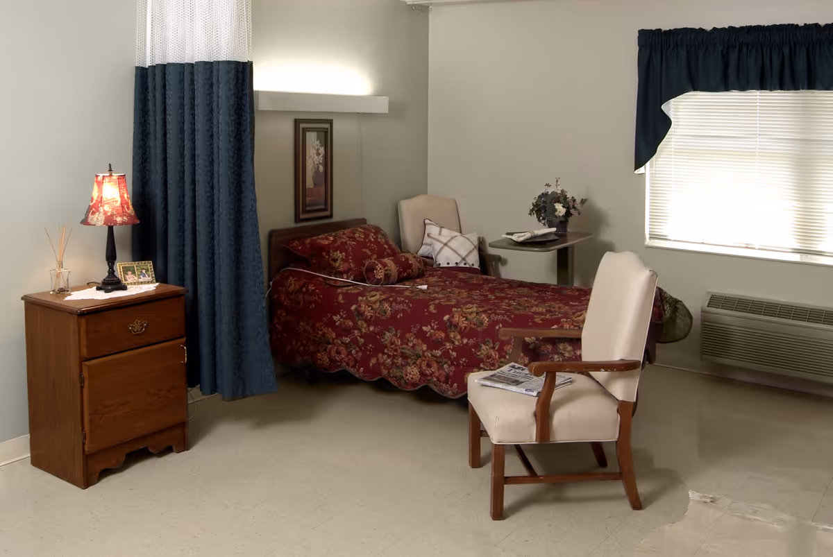 Furnished bedroom with a single bed covered in a red floral bedspread, nightstand with a lamp, and an upholstered chair by a window.