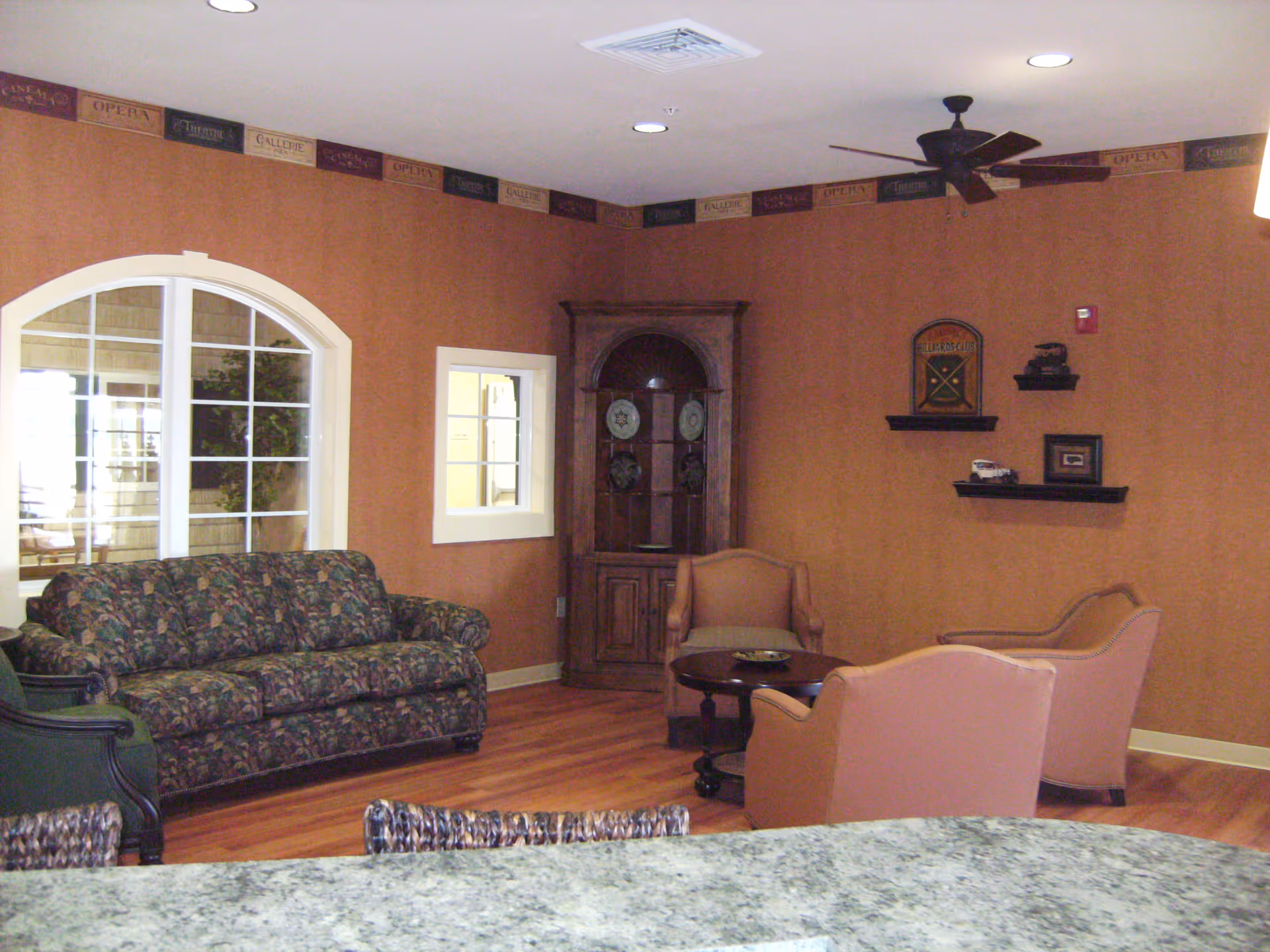 Cozy common sitting area with a floral sofa, several armchairs around a coffee table, wooden floors, an arched interior window, and decorative shelving on a rust-colored wall.
