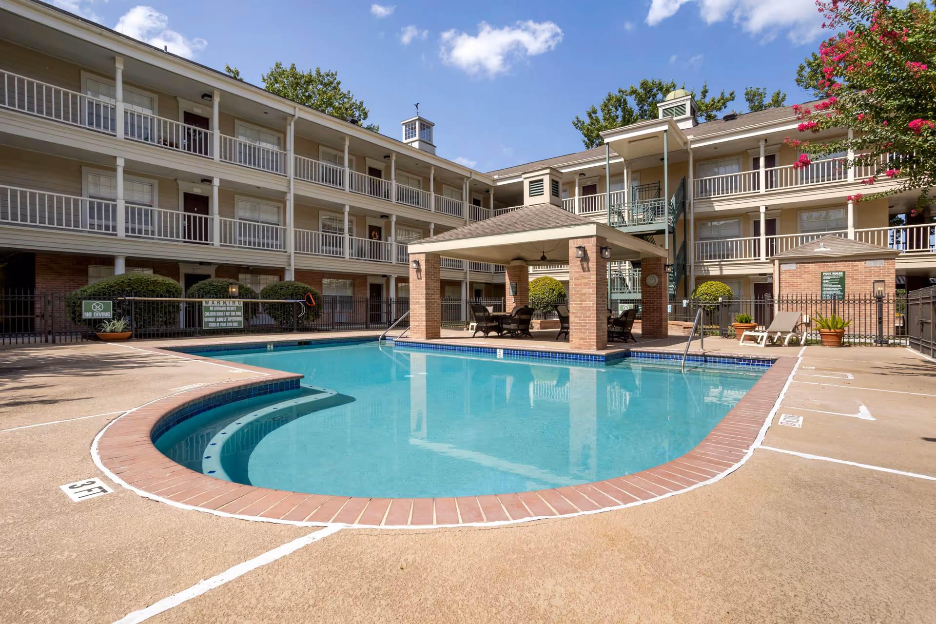 Outdoor swimming pool area at Brookdale Memorial City with a covered seating area in the center of the pool. The pool is surrounded by a concrete deck with lounge chairs and potted plants. A three-story building with balconies and railings encloses the pool area under a clear blue sky.