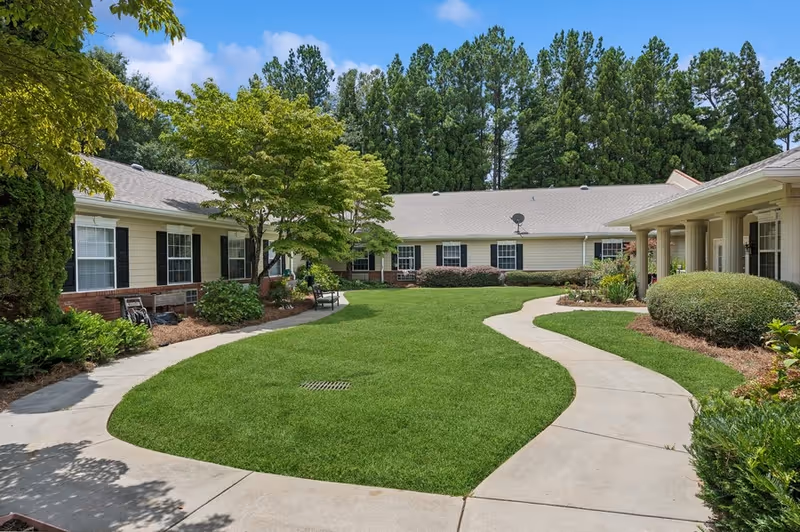 A well-maintained outdoor courtyard area at Gardens of Gainesville featuring a curved concrete walkway surrounding a lush green lawn, with shrubs, trees, and a single-story building with white siding and black shutters in the background under a blue sky.