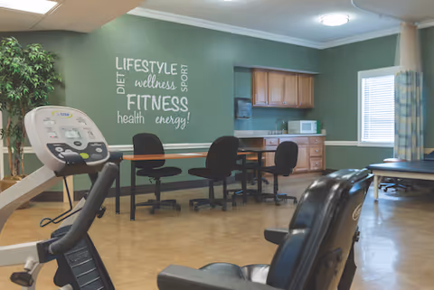 A fitness and wellness room with exercise equipment including a stationary bike and a treadmill. There is a table with four black office chairs against a green wall with words like 'LIFESTYLE', 'FITNESS', 'wellness', 'health', 'energy', 'DIET', and 'SPORT' written on it. The room also has wooden cabinets, a microwave, a window with blinds, and a blue curtain.