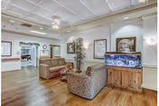 A cozy senior living facility common area with patterned armchairs, a wooden floor, a large fish tank on a wooden stand, framed artwork on the walls, and a ceiling with decorative molding and recessed lighting.
