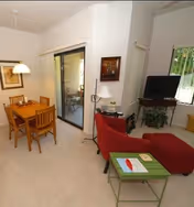 Interior view of a living and dining area in an assisted living facility. The dining area has a wooden table with four chairs and a hanging light fixture above it. Adjacent to the dining area is a sliding glass door leading outside. The living area features a red armchair with an ottoman, a small green table with a book and a card on it, a side table with a lamp, a TV on a stand, and a window with vertical blinds.