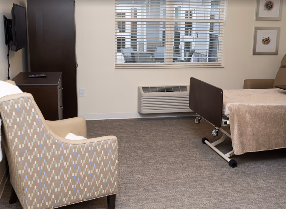 A neutral-toned resident bedroom with a hospital-style bed, patterned armchair, wall-mounted TV, dresser, and window with blinds.