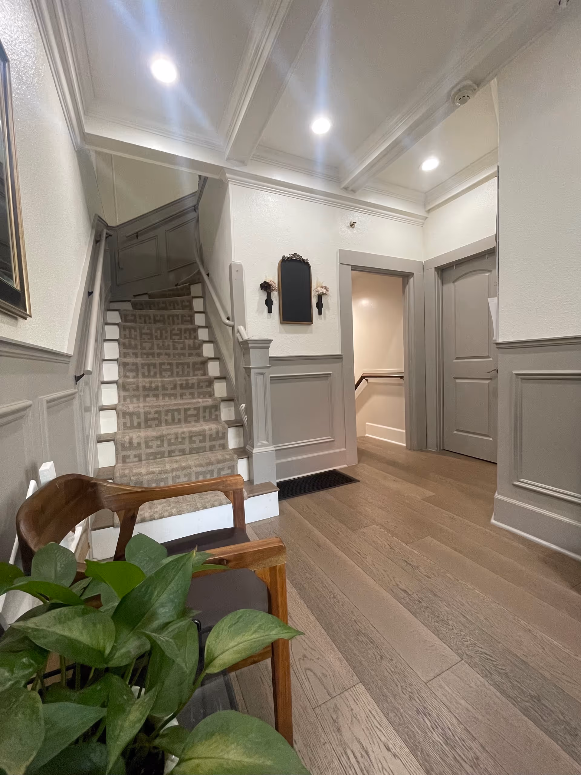 Interior hallway with wooden flooring, a staircase with patterned carpet, two closed doors, a wooden chair with a cushion, and a green leafy plant. The walls are painted white with gray wainscoting, and the ceiling has recessed lighting.