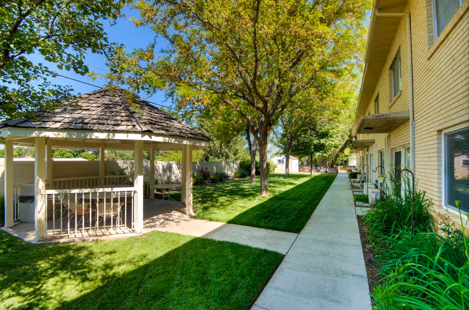 Outdoor area of a senior living facility with a gazebo on the left, green grass, trees, and a concrete walkway alongside a two-story yellow brick building with windows and plants along the building's edge.