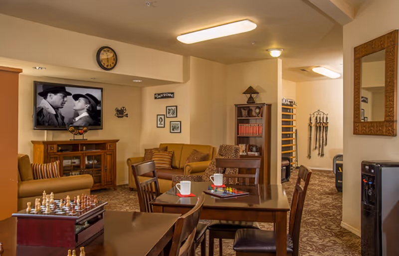 A cozy common area in a senior living facility featuring a wooden table with chairs, a chess set, and board games. In the background, there are comfortable sofas, a wooden cabinet with a TV mounted above showing a black and white movie, framed pictures on the wall, and a clock. The room has warm lighting and a patterned carpet.