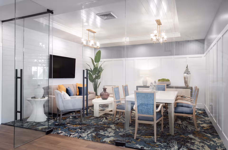 A modern meeting or dining room with glass walls and double glass doors. Inside, there is a white rectangular table surrounded by eight blue cushioned chairs. To the left, there is a beige sofa with colorful pillows, a gray armchair, a white side table with a lamp, and a mounted flat-screen TV on a white shiplap wall. The room features a patterned carpet, two gold chandeliers, a large green plant, and decorative vases on a sideboard against the far wall.