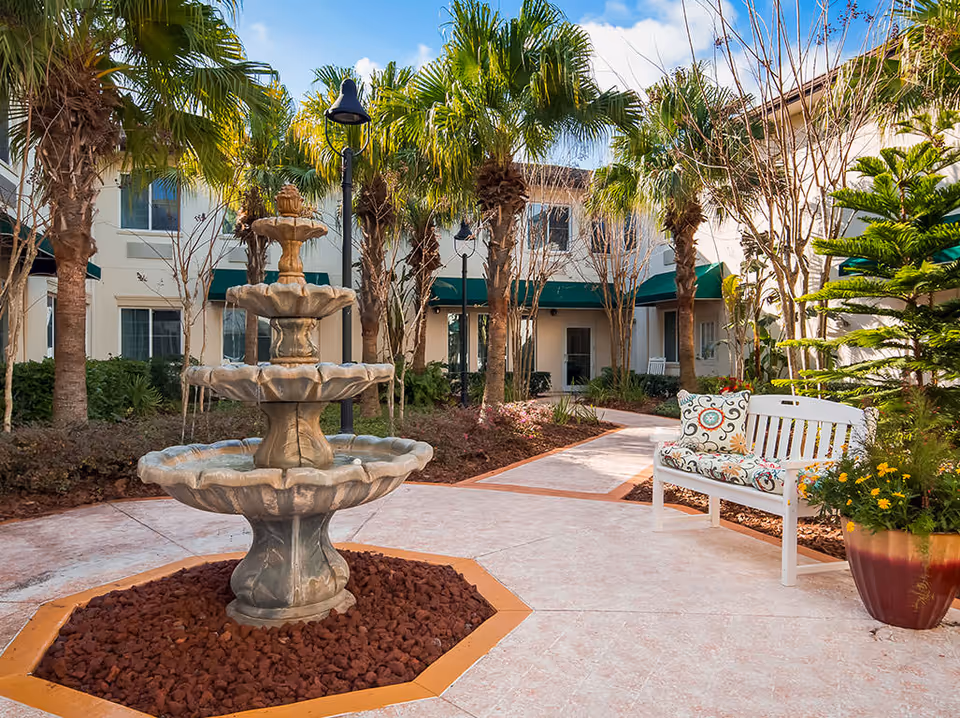 Outdoor courtyard area with a three-tier stone fountain in the center, surrounded by palm trees and landscaped plants. A white bench with colorful patterned cushions is placed on the right side near a large potted plant. The background shows a two-story building with green awnings over the windows and doors.