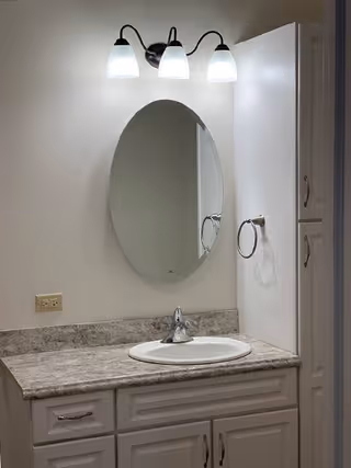 A bathroom vanity with a marble countertop, an oval mirror mounted on the wall, a three-light fixture above the mirror, a sink with a faucet, a towel ring on the right wall, and white cabinetry below and to the side of the countertop.